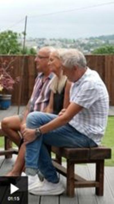 Three people sit on a bench, side on to the camera. Furthest from the camera is an older man, balding and wearing glaases, a striped shirt and shorts. Next to him sits a woman with blonde hair wearing a black top. Closest to the camera is a man with grey hair, wearing a grey and white striped shirt, blue jeans and white trainers. They are in a garden, with a lawn and fence in the background.