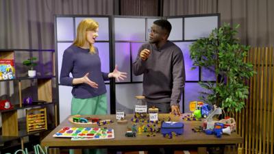 De-Graft meets Coletta Smith to find out about the Autumn Budget. They are standing behind a table with lots of toys and props on it.