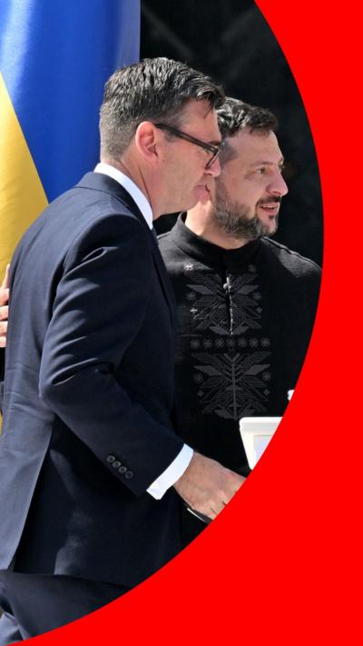 Ukraine President Volodymyr Zelensky has his arm around UK Prime Minister Andy Burnham's shoulder. There is a Ukrainian flag behind them.