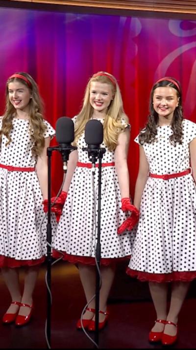 Three women dressed in white dresses with black spots and red belts, with red shoes. They are on a stage singing into a microphone.