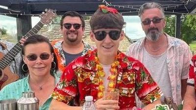 Bailey Chadwick in the centre of the image wearing a red Hawaiian shirt and sunglasses. He is stood at a bar with other employees at the pub wearing similar outfits.