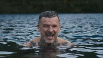 A tattooed man smiling, he is submerged in water up to his shoulders.