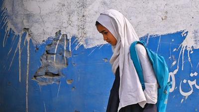 girl wearing a headscarf and a backpack walks beside a wall with her head down