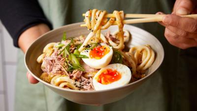 A bowl of udon noodles with sliced boiled egg and tuna on the top, with someone holding chopsticks holding some of the noodles.