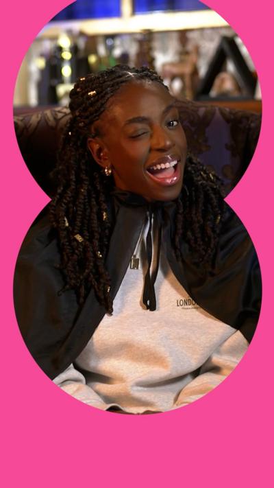 Cat Burns winks while sat in an armchair wearing a dark green cloak over a grey sweater. She has long black braids