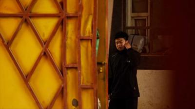 A man in a black jacket holds his ear and watches on as the camera films