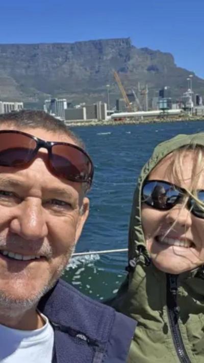 Man with moustache and trimmed beard wearing sunglasses on his brow smiling at the camera. He is wearing a fleece and a white t-shirt. Sitting next to him is a girl with her hair flying in the wind. She has a hood up on a waterproof she is wearing. She is smiling and they are both on a yacht. Behind them is Table Mountain.