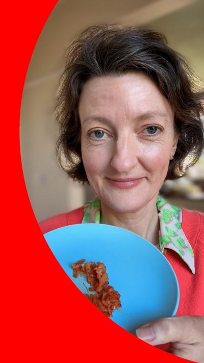 A woman with brown hair holds up a blue plate holding kimchi. She is wearing a dark pink jumper and a green-collared shirt.