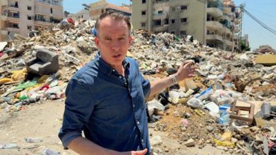 John Sudworth points towards rubble, the remains of a building, in Dahieh