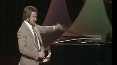 Richard Stilgoe sits at a piano.