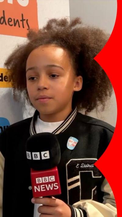A boy with brown hair and a letter man style jacket holds a BBC News branded microphone