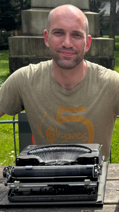 Patrick, a man with a shaved head and stubbled facial hair, smiles sitting behind his typewriter on a wooden picnic bench in Botanic. 