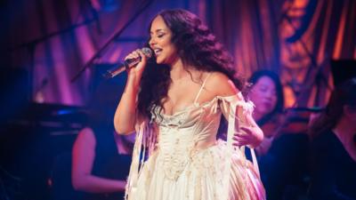 Jade sings on stage. She has long wavy black hair and wears a ruffled white dress. Behind her are members of an orchestra