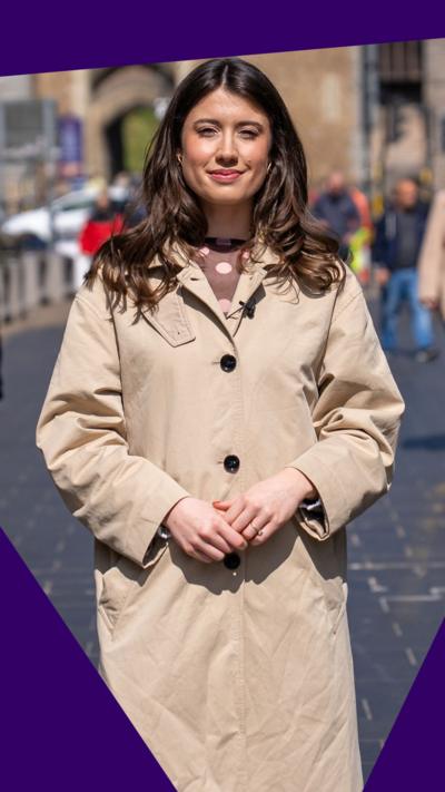Meleri Williams, who has long brown hair and is wearing a long beige coat buttoned up. The entrance to a castle and people are in the background