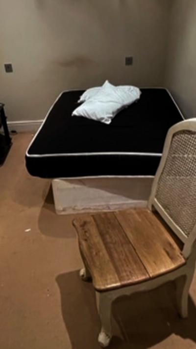 A wooden chair is in front of a black mattress with two white pillows on top of it. The items are in a room with brown carpet and beige walls