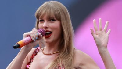 American popstar Taylor Swift waving. She is holding a pink an orange micrphone.