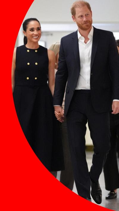 Meghan wearing black dress with golden buttons and holding hands with Harry who is wearing a white shirt and black suit