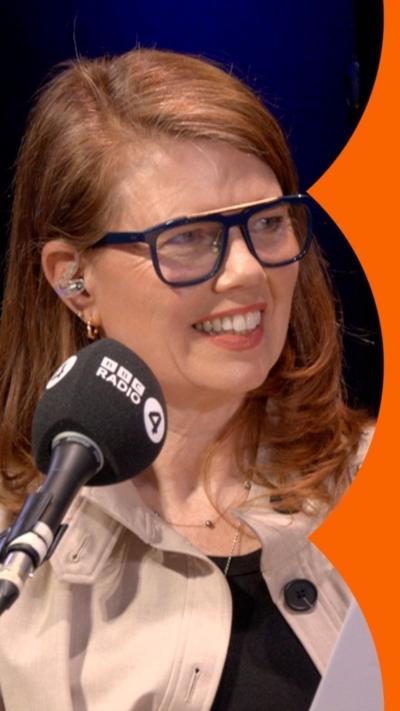 A woman with auburn hair and glasses sits behind a microphone. She is smiling at someone on her left.