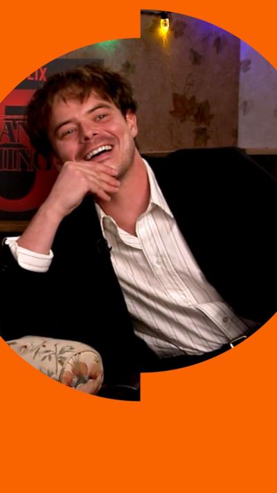 Charlie Heaton in a black suit and white shirt, smiling, leaning on his arm.