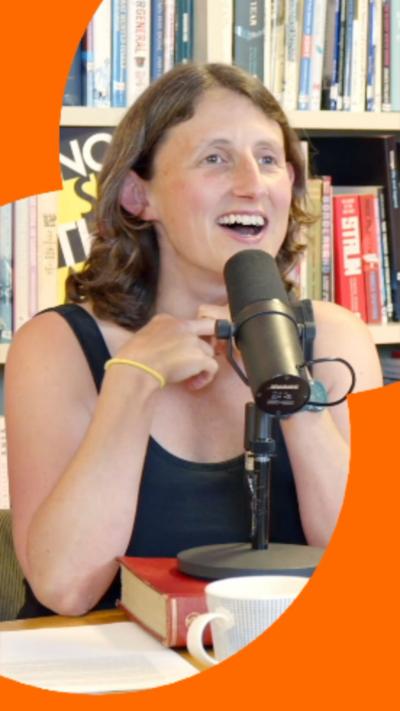 A woman with long brown hair wearing a navy blue top smiles with a microphone in front of her.