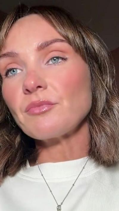 A woman with mid length dark hair looks into the camera. She is wearing a white round neck top and a thin necklace.