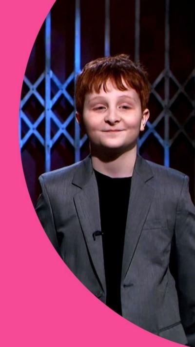 A pre-teen boy smiling, wearing a grey blazer and black t-shirt, with dark red hair.