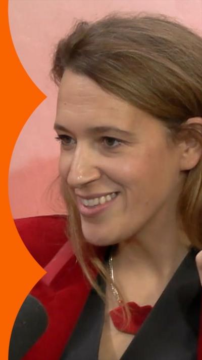 A woman, Dr Filby, has long brown hair and wears a black top with a red jacket