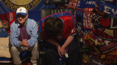 A man sits next to a younger boy in a wheelchair with a wall of football scarves behind 