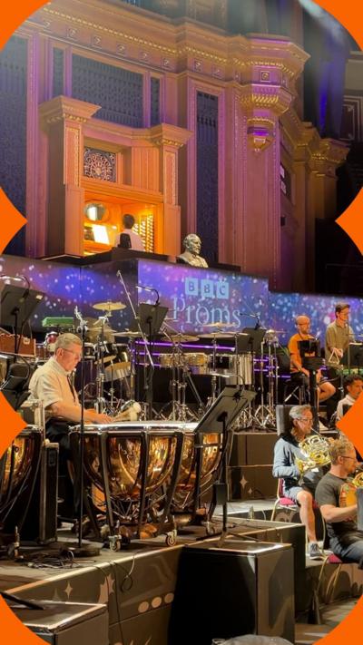 Members of the orchestra sit in front of a sign saying BBC Proms. In the foreground, a man sits playing kettle drums.