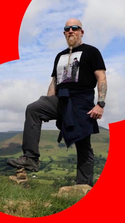 A man with tattoos, sunglasses and a plaited beard, with one foot on a stone, and the Peak District hills behind him.