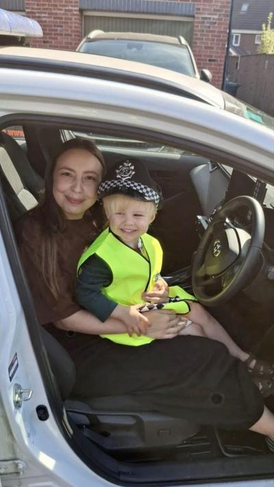 A young boy dressed in a police uniform sat on a woman's lap in a car seat