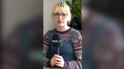 A teenage girl holds a 5 Live mic. She has short blonde hair and wears a purple patterned jumper and denim dungarees