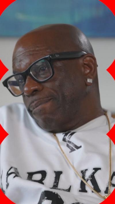 A man with a gold chain and black glasses speaks into the camera.