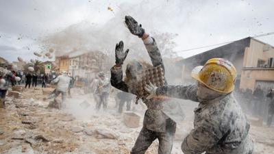 A man in a yellow helmet dressed in old-fashioned military gear splats another person with a pallet of eggs.