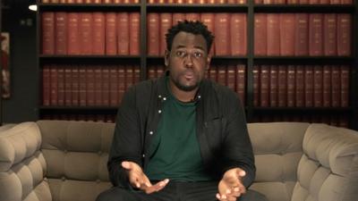 Man looking confused sitting in front of books. He has a black shirt on over a dark green t-shirt.