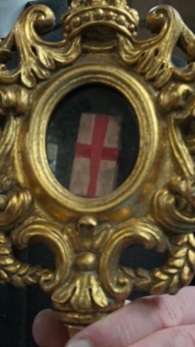 Close up image of vicar's hand. The vicar is wearing balck clothing with a white collar and is holding an intricate gold painted reliquary which houses a small wooden box which is tied by a red ribbon making the shape of a red cross.