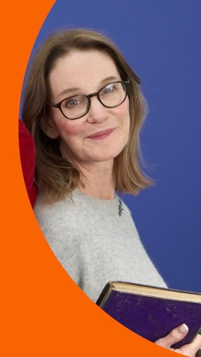 Susie Dent wears a grey jumper and holds a book