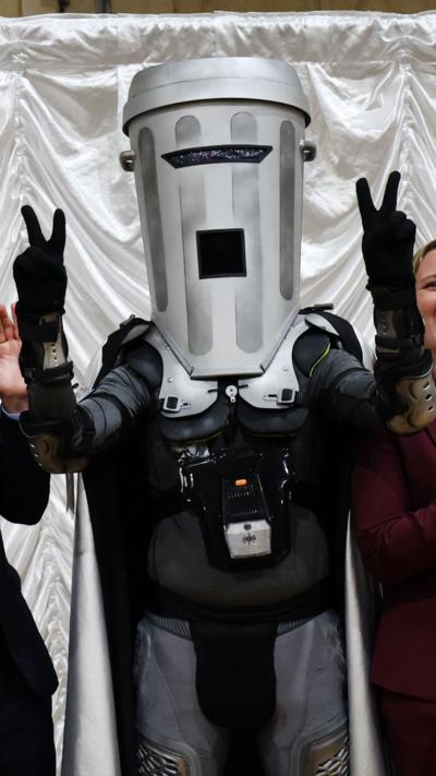 Man wearing grey bin mask over his head lifting two arms and holding his fingers in peace signs