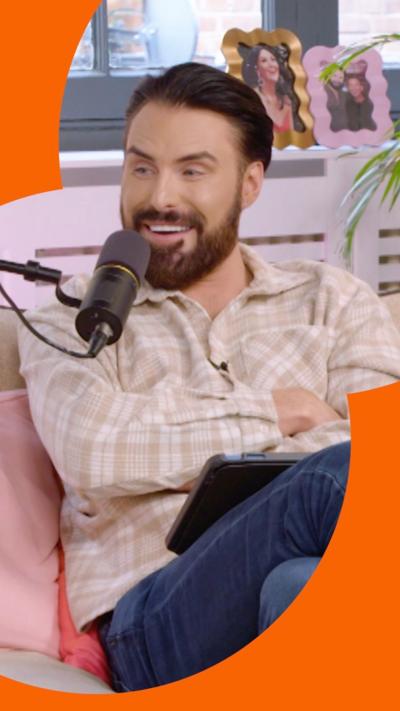 Rylan Clark-Neal sits on a sofa smiling - he has short black hair and a beard