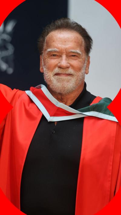 Arnold Schwarzenegger smiles at the camera. He has short, dark hair and a short, white beard. He wears a red graduation gown.