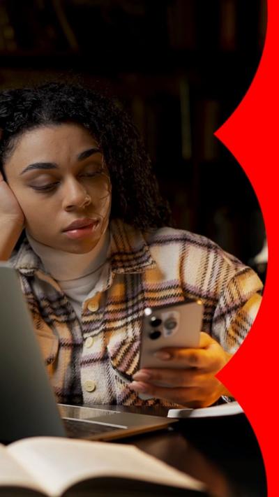 A woman checks her phone in the library. She is wearing a checkered shirt.