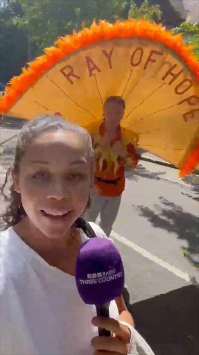 Female reporter with dark curly hair tied back wearing a white top, standing in front of a man with a Ray Of Hope giant fan on his back in a carnival parade