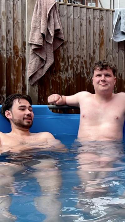 Two men sat in a blue paddling pool, in front of a wooden fence that's been splashed with water and has a brown towel thrown over it.