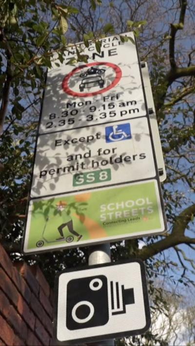 A street sign setting out the details of parking restrictions. The signpost also shows that it is in a School Streets area and features an image of a camera