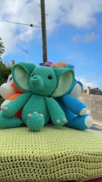 A crochet postbox topper with a green elephant facing towards the camera and blue and orange animals facing away. The sky is blue with clouds.