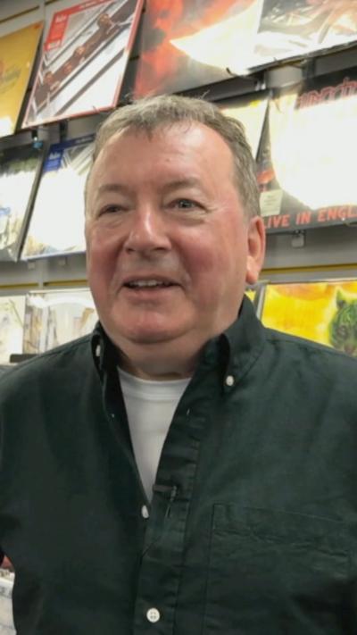 Raymond Stewart is standing in front of several rows of vinyl records. He is smiling and is wearing a grey shirt with a white T-shirt underneath. 
