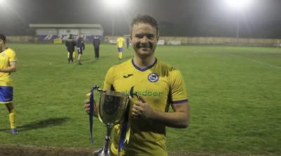 Joe Medcalf wearing football kit holding a trophy on a football pitch