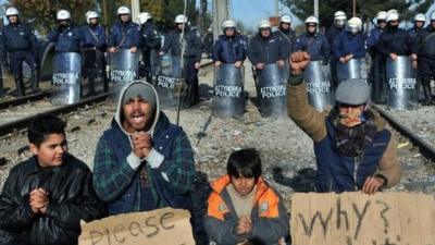 Greek police officers stand guard behind two men and two children demonstrating between railway tracks as they wait with other migrants and refugees to cross the Greek-Macedonian border near Idomeni on 7 December