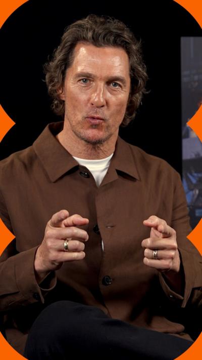 Matthew McConaughey gestures towards the camera during an interview. He wears a brown collared jacket