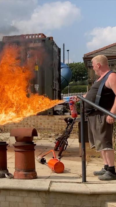 Man in a black vest and dark shorts holds a fire extinguisher that has been converted into a flame thrower.